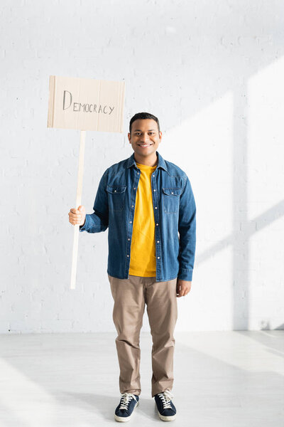smiling indian man holding placard with democracy lettering against white brick wall