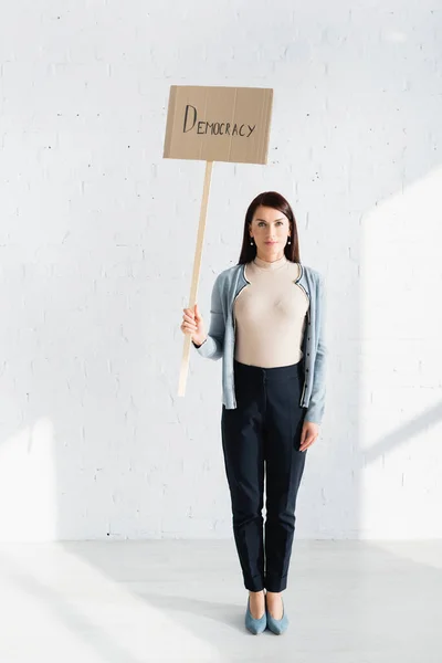 Allvarlig Kvinna Håller Plakat Med Demokrati Inskription Mot Vit Tegelvägg — Stockfoto