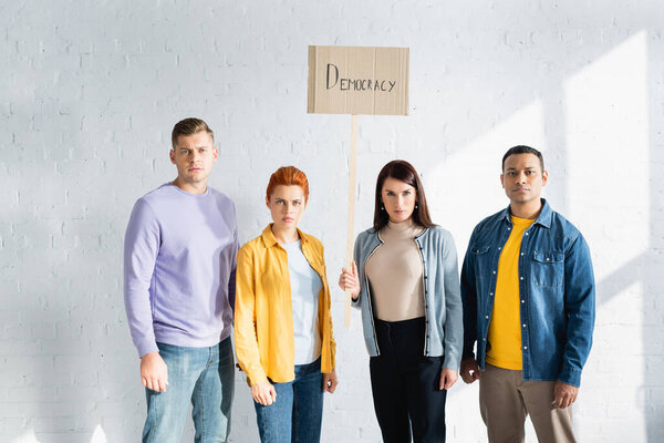 serious multicultural people looking at camera while holding placard with democracy lettering