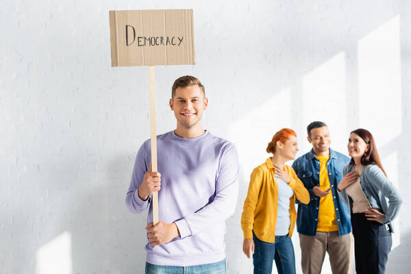 happy man holding placard with democracy inscription while multicultural like-minded people talking on blurred background