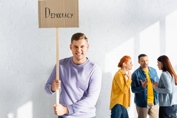 angry man grimacing while holding placard with democracy lettering near multicultural people talking on blurred background