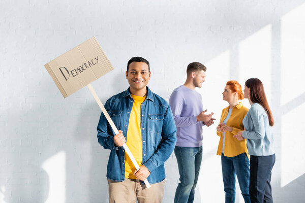 happy indian man holding placard with democracy lettering while multicultural like-minded people talking on blurred background