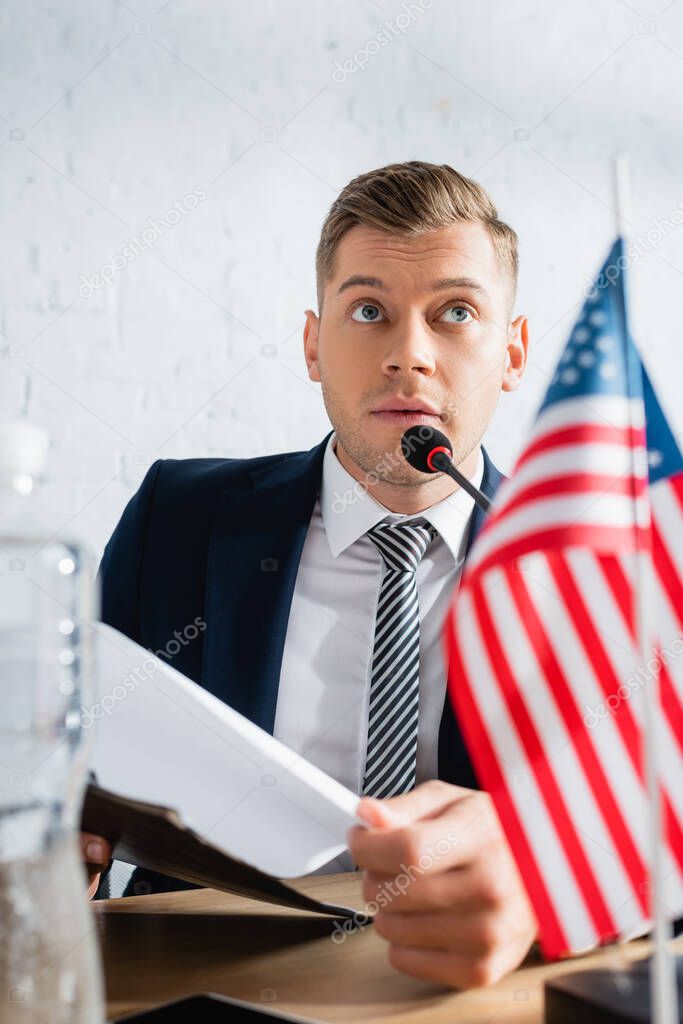 Confident member of political party holding clipboard, while speaking in microphone at table with usa flag on blurred foreground