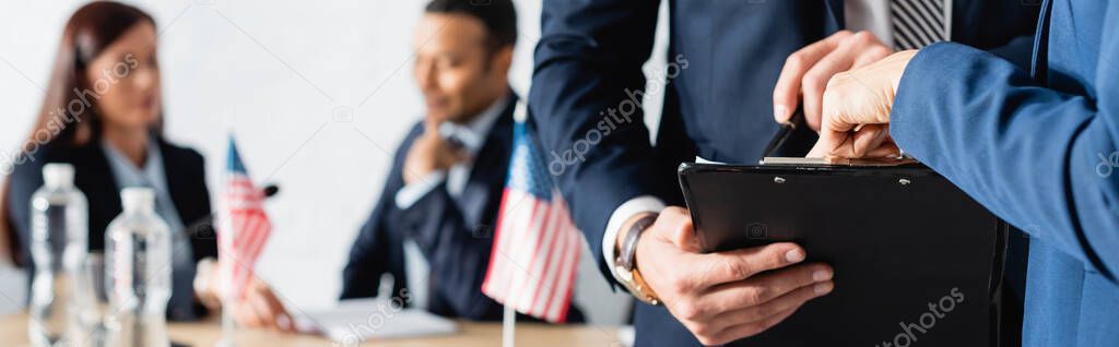 politician pointing with finger at clipboard near colleagues talking on blurred background, banner