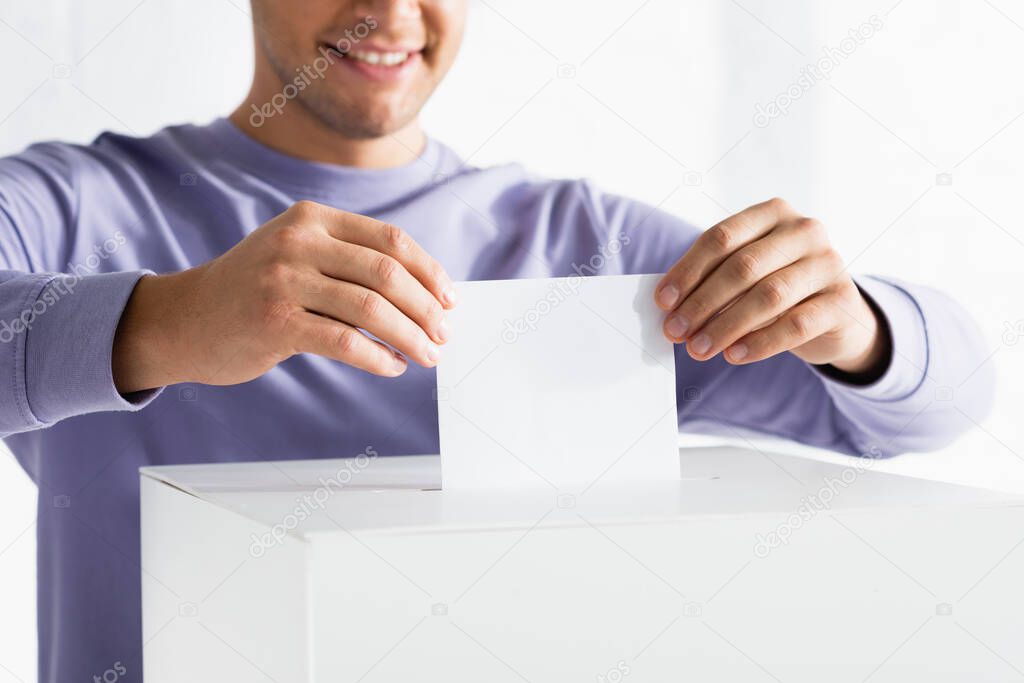 cropped view of smiling man inserting ballot into polling box on blurred background