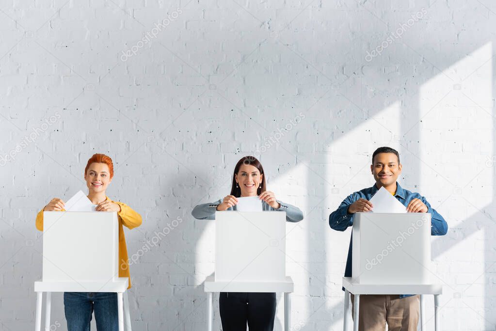 multicultural voters inserting ballots into polling boxes against white brick wall