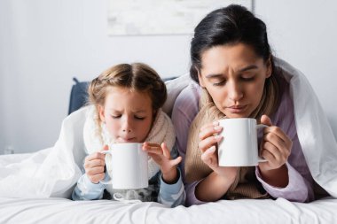 Hastalıklı anne ve kızı yatakta yatarken sıcak çay üflüyorlar.