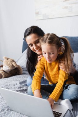 Mutlu bir kadın yatak odasında dizüstü bilgisayar kullanarak kızını kucaklıyor.