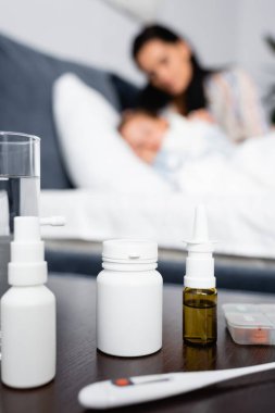 bottles with medicines on bedside table near sick child lying in bed with mother on blurred background clipart