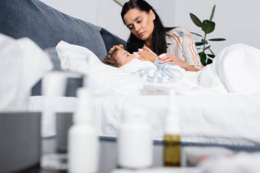 Hasta kızın yanında yatan, kağıt peçeteye hapşıran bir kadın ve bulanık ön planda ilaçlarla birlikte komodinin yanında yatan bir kadın.