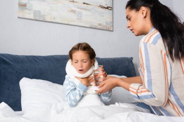 Endişeli anne yatakta öksüren hasta kıza bir bardak su veriyor.