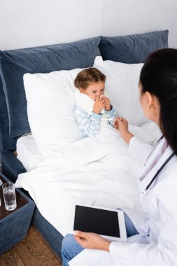Pediyatrist hasta çocuğa termometre veriyor. Yatakta yatıyor, ön plan bulanık.