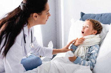 Pediyatrist yatakta yatan hasta kızın boğazını muayene ediyor.