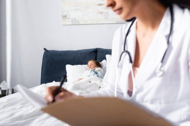 Hasta çocuk, çocuk doktoru önplanda bulanık teşhis yazarken yatakta yatıyor.