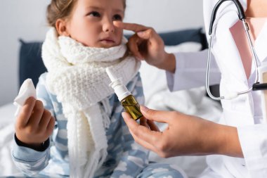 Doktor, bulanık arka planda burun spreyi tutarken hastalıklı kızın burnunu işaret ediyor.