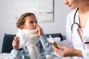 Hasta çocuk burnuna işaret ediyor ve doktor burun spreyi tutarken kağıt peçete tutuyor.