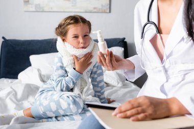 Doktor, boğaz spreyini boynunda sıcak atkısı olan, ön planda bulanıklık olan hasta kıza gösteriyor.