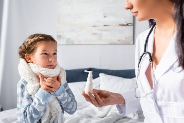 Pediyatrist, boynuna sıcak atkı takmış hasta kıza boğaz spreyi sıkar.