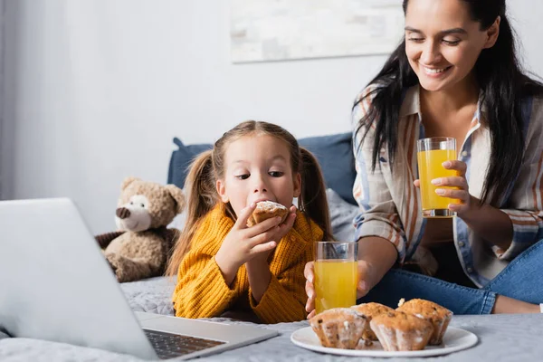 ラップトップで映画を見ながら陽気な母親の近くでマフィンを食べる女の子 — ストック写真