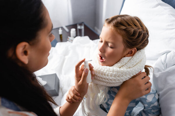 mother applying spray on sore throat of sick daughter on blurred foreground