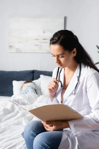 serious doctor looking at outpatient card while diseased child sleeping in bed on blurred background