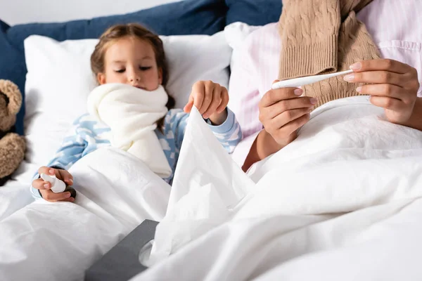 diseased girl with nasal spray and mother with thermometer lying in bed