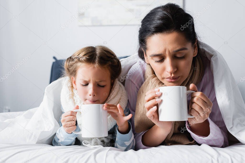diseased mother and daughter blowing on hot tea while lying in bed