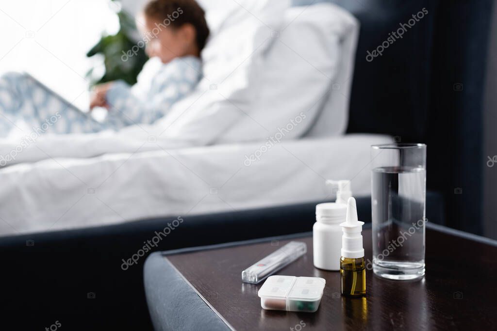 bedside table with glass of water and medicines near sick girl lying in bed on blurred background