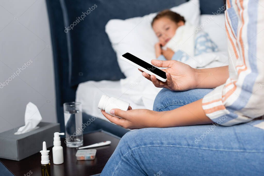 mother holding bottle of pills and mobile phone near sick daughter lying in bed