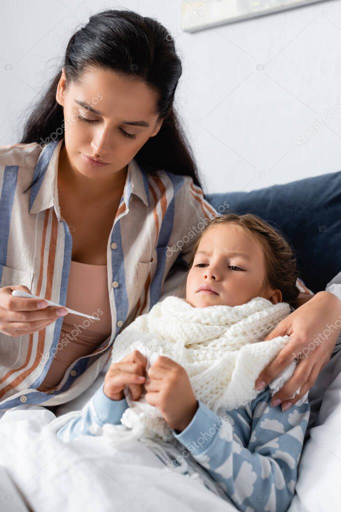 woman looking at thermometer near ill daughter lying in bed