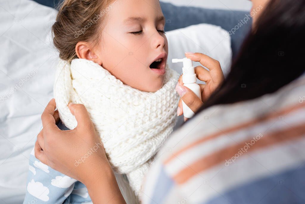 sick child with closed eyes and open mouth near mother applying spray on sore throat on blurred foreground