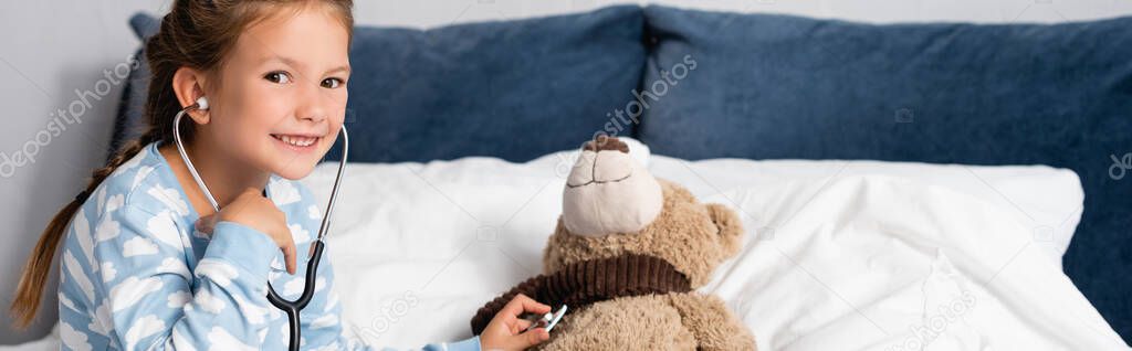 cheerful girl looking at camera while examining teddy bear with stethoscope, banner