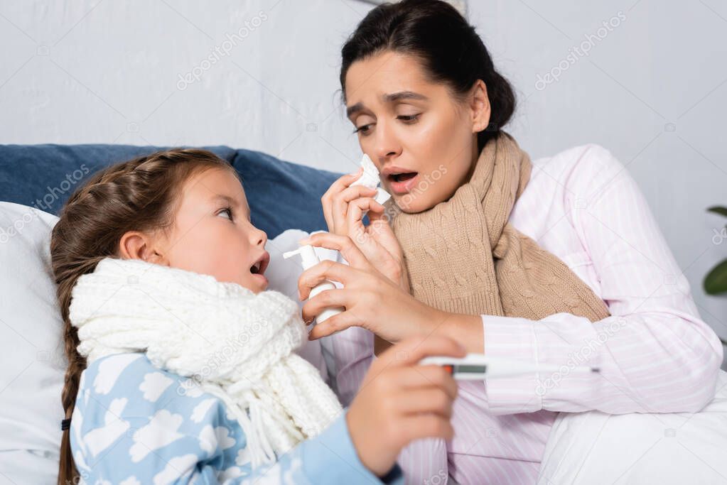 ill woman with runny nose holding throat spray near diseased girl with thermometer on blurred foreground