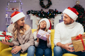 radostné rodiče v Santa klobouky držení dárky a při pohledu na dítě v zdobeném obývacím pokoji na Vánoce 