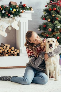 Kazaklı mutlu kadın yerde oturuyor ve yılbaşı ağacının yanında Labrador 'a sarılıyor.