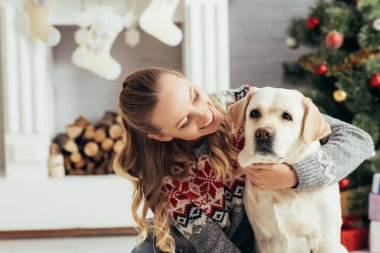 Kazaklı mutlu kadın, yılbaşı ağacı ve şöminesinin yanında Labrador 'a bakıyor.