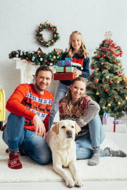 Süveter giyen mutlu ebeveynler kızlarının yanında Labrador 'a sarılıyorlar hediyeler ve süslü Noel ağacı ile.