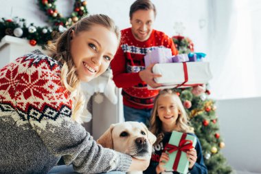 Mutlu bir kadının, kızının ve kocasının Noel hediyeleriyle köpeğini kucaklamasının seçici odağı. 