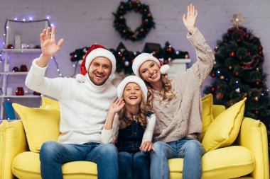 Noel Baba şapkalı mutlu aile kameraya bakıyor ve dekore edilmiş oturma odasında el sallıyor. 