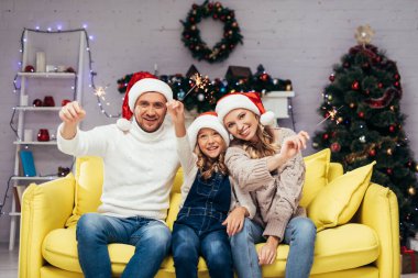 Noel Baba şapkalı mutlu aile dekore edilmiş oturma odasında maytap tutuyor. 