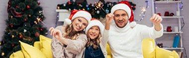 happy family in santa hats holding sparklers in decorated living room, banner clipart