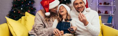 Mutlu anne ve kız Noel Baba şapkaları içinde Patlamış mısır ve afiş yiyen adamın yanında gülüyorlar.