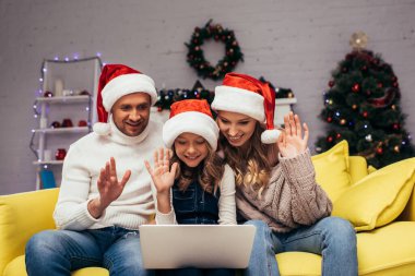 Noel Baba şapkalı neşeli bir aile dekore edilmiş oturma odasında görüntülü sohbet ederken el sallıyor. 