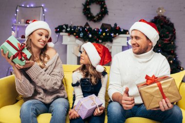 Noel Baba şapkalı neşeli aile Noel 'de dekore edilmiş oturma odasında hediyeler tutuyor. 