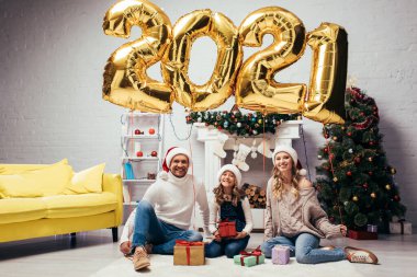Noel Baba şapkalı neşeli aile hediyelerin yanında oturuyor ve üzerinde 2021 numara olan parlak balonlar. 