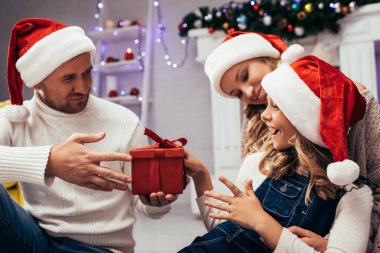 Noel Baba şapkalı baba, karısının yanındaki mutlu kızına hediye veriyor.