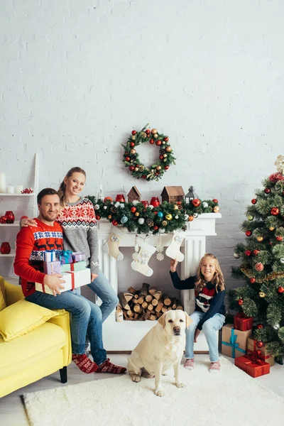 happy parents holding gifts near daughter with dog and decorated christmas tree