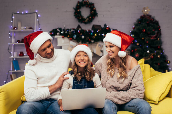 счастливый малыш в шляпе санта с ноутбуком рядом с родителями в украшенной гостиной 