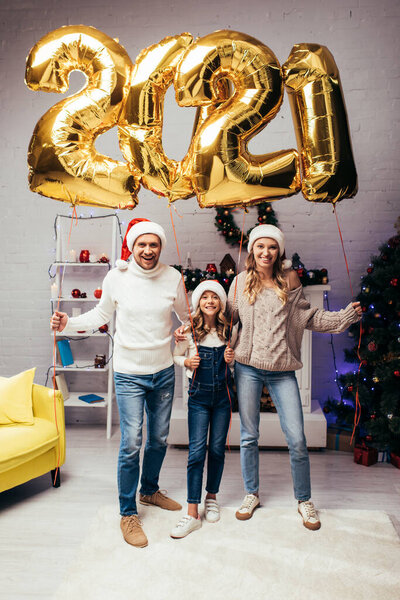 happy family in santa hats standing near presents and holding shiny balloons with 2021 numbers 