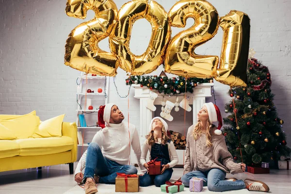 戴着桑塔帽的家庭惊奇地坐在礼物旁边 手里拿着2021年数字的闪亮气球 — 图库照片